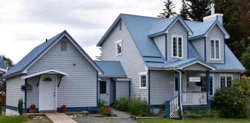A two-story house with a blue metal roof, white siding, and a small porch, surrounded by greenery.