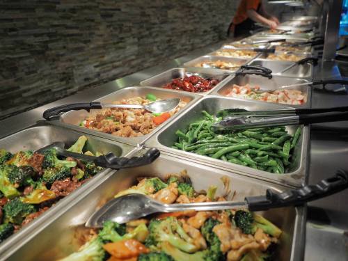 A buffet with various dishes, including vegetables and meats, in metal trays with serving tongs.