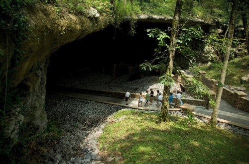 Visitors walk along a path near a cave entrance surrounded by lush greenery and rocky terrain.