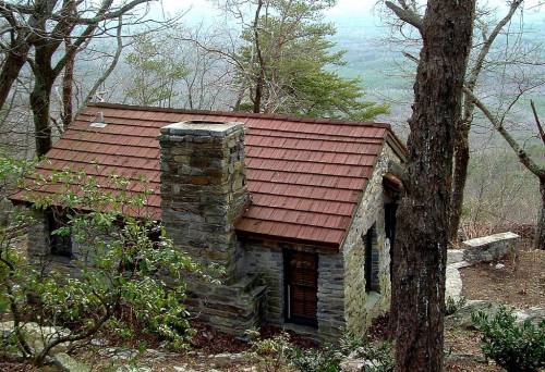 A small stone cabin with a red roof nestled among trees on a hillside, overlooking a misty valley.