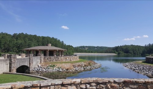 A serene lake view with a stone pavilion, surrounded by trees and a dam under a clear blue sky.