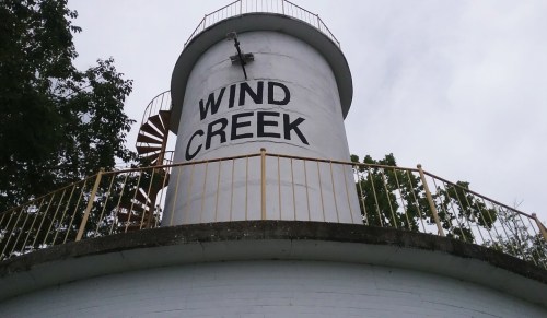 A white water tower labeled "WIND CREEK" with a spiral staircase and a railing, surrounded by trees under a cloudy sky.