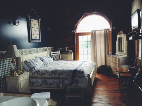 Cozy bedroom with a large bed, wooden floors, and a window letting in natural light. Decor includes a lamp and a small table.