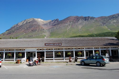 A rustic motor inn with flower baskets, set against a backdrop of mountains under a clear blue sky.