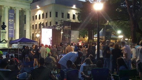 A crowd gathers at an outdoor event with a stage, bright lights, and tents in a park setting at dusk.