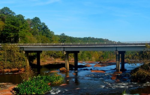 A bridge spans a river surrounded by lush greenery and rocky banks under a clear blue sky.