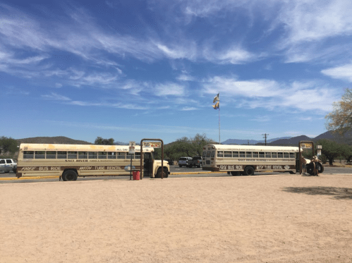 Two vintage yellow school buses parked on a sandy area under a blue sky with scattered clouds.