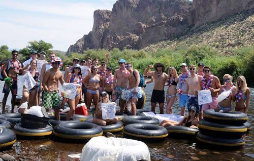 A large group of people in swimsuits enjoying a summer day on inflatable tubes in a river, surrounded by rocky hills.