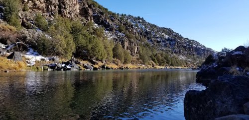 A serene river flows between rocky banks, surrounded by lush greenery and snow-capped mountains under a clear blue sky.