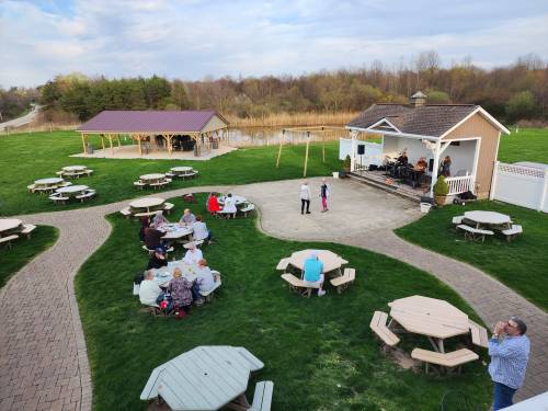 Outdoor venue with picnic tables, a stage with a band, and people enjoying a gathering near a pond.