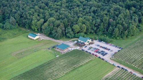 Aerial view of a farm with green fields, buildings, and a large parking area filled with cars. Surrounding forest in the background.