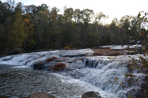 A serene waterfall cascades over rocks, surrounded by lush trees and a tranquil river under a soft sunset.