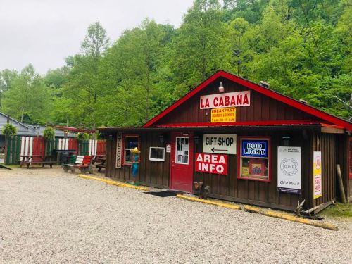 A rustic wooden building with a red roof, featuring signs for a gift shop and breakfast, surrounded by greenery.