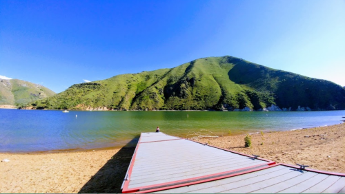 A serene lake with a wooden dock extending into the water, surrounded by lush green hills under a clear blue sky.