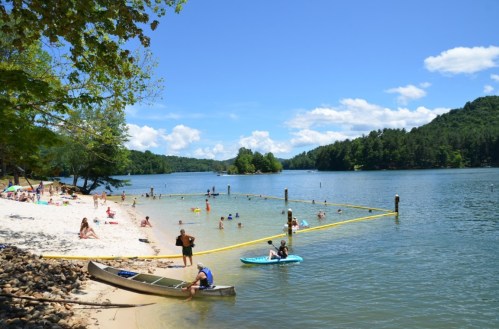 A sunny lakeside scene with people swimming, a sandy beach, and kayaks near a yellow floating barrier.