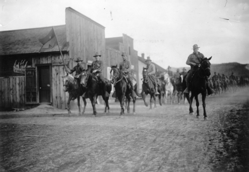 A line of mounted soldiers riding down a dirt street, flanked by wooden buildings in a historical setting.