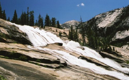 A cascading waterfall flows over smooth rocks, surrounded by tall trees and mountains under a clear blue sky.
