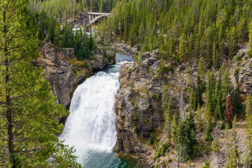 A waterfall cascades down rocky cliffs, surrounded by lush green trees and a bridge in the background.
