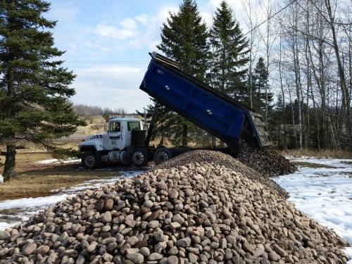 A dump truck unloading a large pile of rocks in a snowy yard, surrounded by trees and a clear sky.