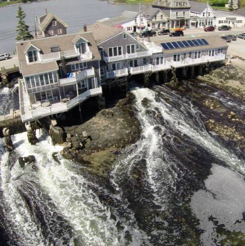 A coastal building on stilts overlooking turbulent waves and rocky shoreline, with nearby houses and a calm harbor in the background.
