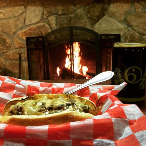 A hot dog topped with melted cheese sits on a red checkered paper, with a cozy fireplace in the background.