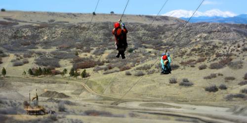 Two people ziplining over a hilly landscape with sparse vegetation and snow-capped mountains in the background.