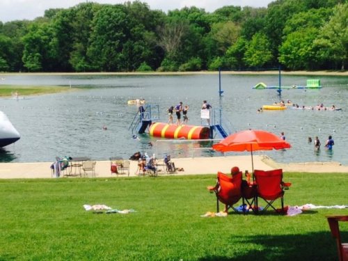 A sunny lakeside scene with people swimming, a colorful inflatable, and red chairs under an umbrella on the grass.