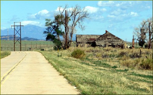 A weathered barn beside a dirt road, with distant mountains and trees under a partly cloudy sky.
