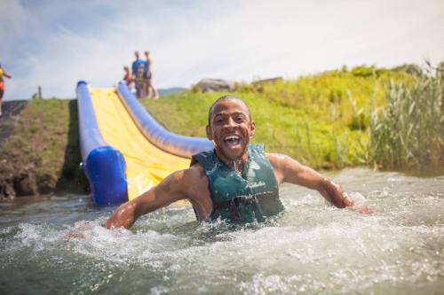 A smiling boy in a life jacket splashes in water near a yellow slide, with people enjoying the background.