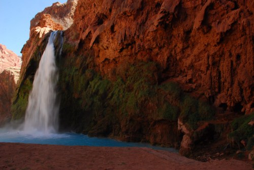 A stunning waterfall cascades over red rock cliffs into a turquoise pool, surrounded by lush greenery.