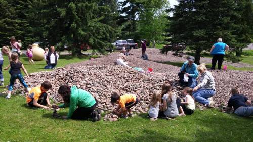 A group of children and adults are digging and playing in a large pile of rocks in a grassy area surrounded by trees.