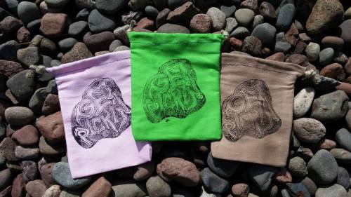 Three colorful fabric bags in pink, green, and brown, each featuring a unique abstract design, placed on a bed of pebbles.