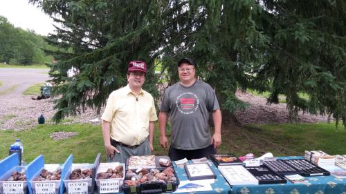 Two men stand behind a table filled with various items for sale, set up outdoors under a large tree.
