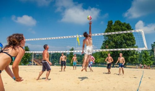 A group of people playing beach volleyball on a sunny day, with one player jumping to hit the ball over the net.