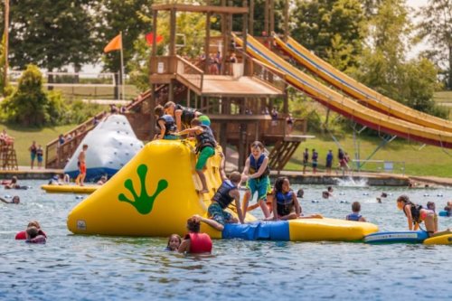 Children play on a colorful inflatable structure in a lake, with water slides and other kids swimming nearby.