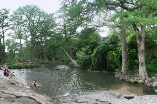 A serene river scene with people swimming, surrounded by lush greenery and tall trees. Rocky banks frame the water.