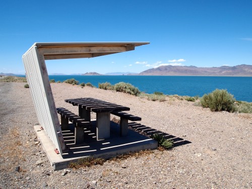 A shaded picnic table by a lake, surrounded by shrubs and mountains under a clear blue sky.