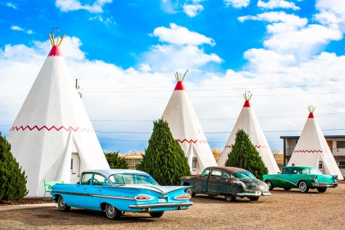 Colorful vintage cars parked in front of white teepee-shaped buildings under a blue sky with fluffy clouds.