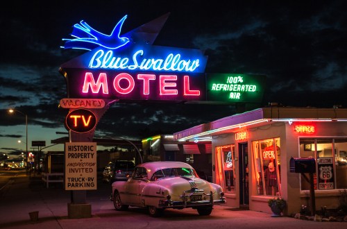 Neon sign of Blue Swallow Motel at dusk, featuring a classic car parked outside and a retro vibe.