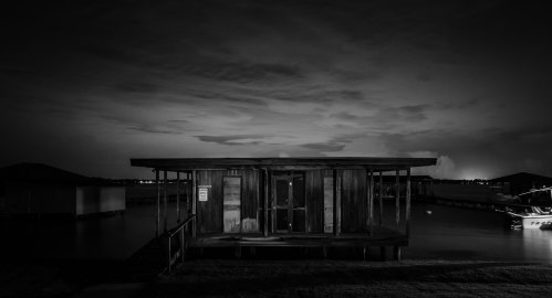 A dark, moody image of a wooden cabin by the water, surrounded by clouds and distant lights at night.