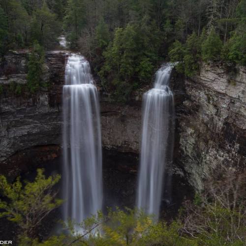 Two cascading waterfalls surrounded by lush green trees and rocky cliffs in a serene natural setting.