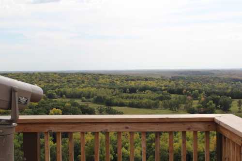 A scenic view from a wooden deck overlooking lush green hills and trees under a partly cloudy sky.