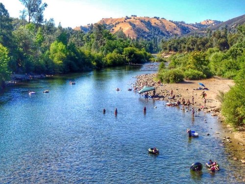 A scenic river with people swimming and relaxing on the shore, surrounded by lush greenery and hills in the background.