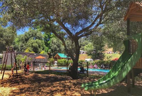 A playground with a green slide and swing set, surrounded by trees, near a pool with people enjoying the sunny day.