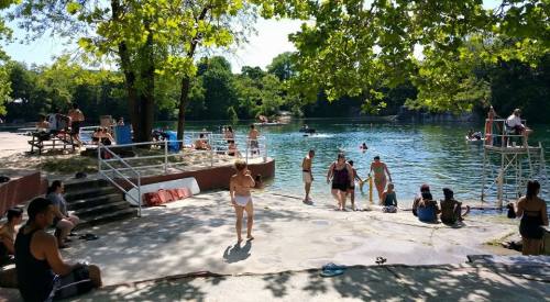 A sunny lakeside scene with people swimming, lounging, and enjoying the water surrounded by trees.
