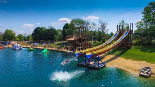 A vibrant water park scene featuring a large slide, swimmers, and inflatable attractions by a lake under a clear blue sky.