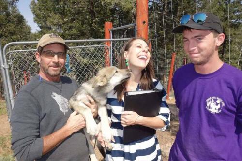 A man holds a puppy while a woman laughs and a young man looks on, all in a fenced outdoor area.