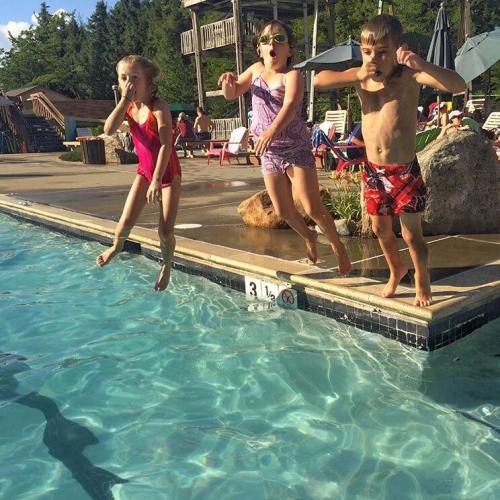 Three children are jumping into a pool on a sunny day, with lounge chairs and trees in the background.