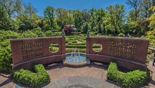 A serene garden with circular brick walls, a fountain, and lush greenery, surrounded by trees and a gazebo in the background.