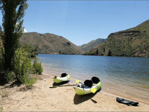 Two kayaks on a sandy shore by a calm lake, surrounded by green hills under a clear blue sky.